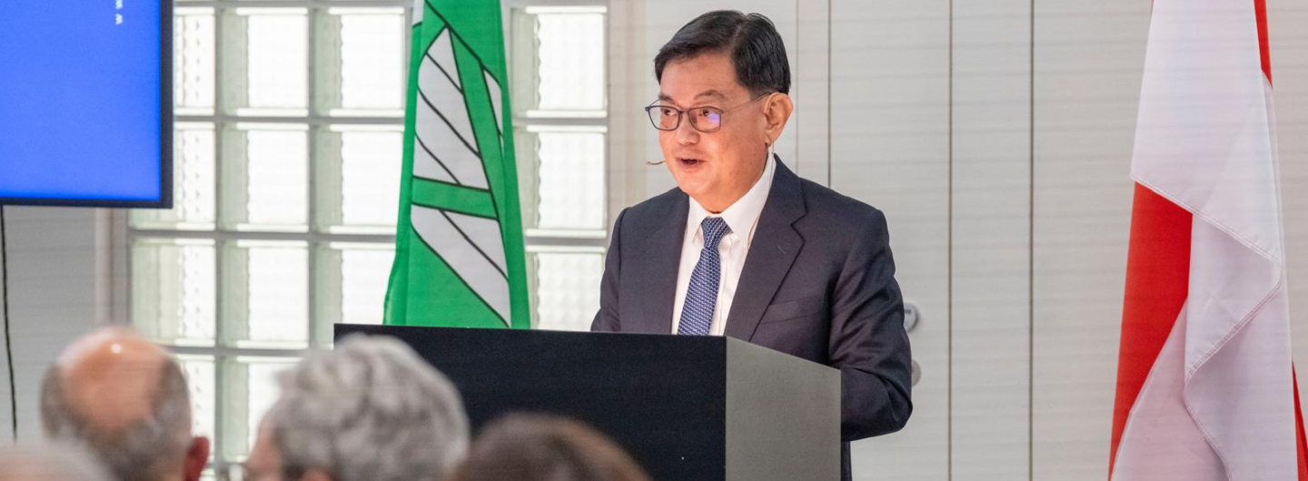 Man at podium with flags, wearing suit, glasses and tie, speaking to audience.
